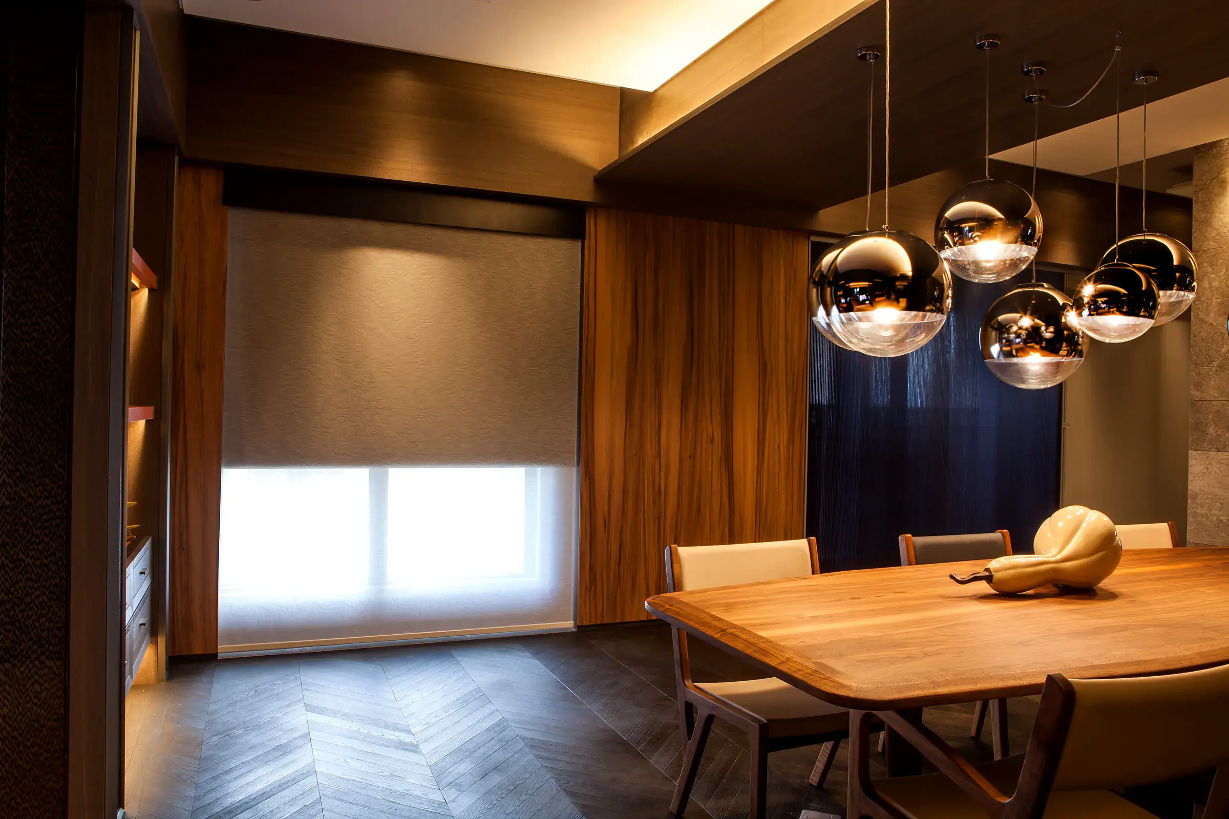 Dual roller shades in a warmly lit dining room: a brown patterned roller shade partly lowered in front of a separate sheer white roller shade, beside timber wall panels, a walnut table and mirrored pendant lights.