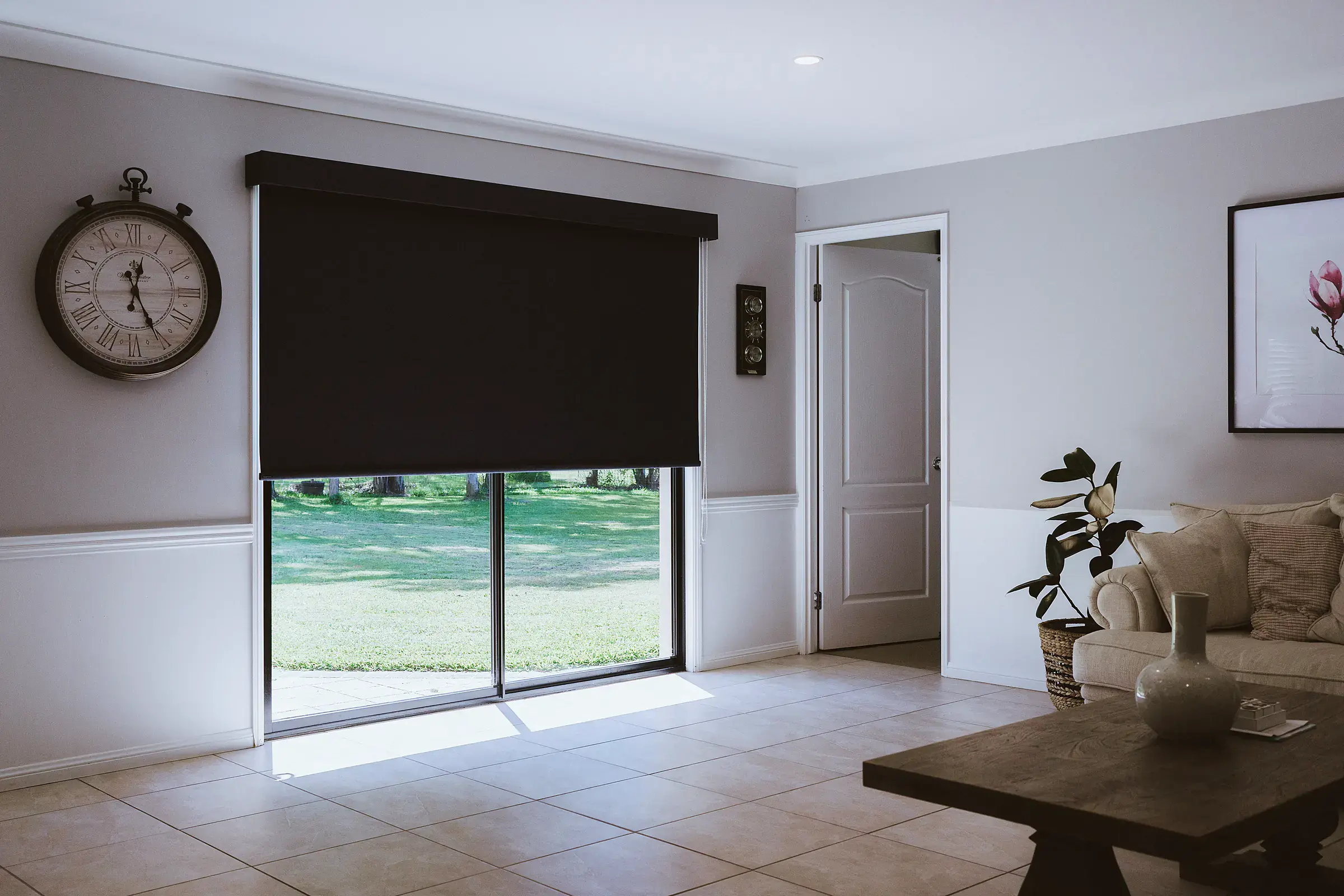 Black roller shade with a matching pelmet, partly lowered over sliding glass doors in a living room that opens onto a sunny garden lawn