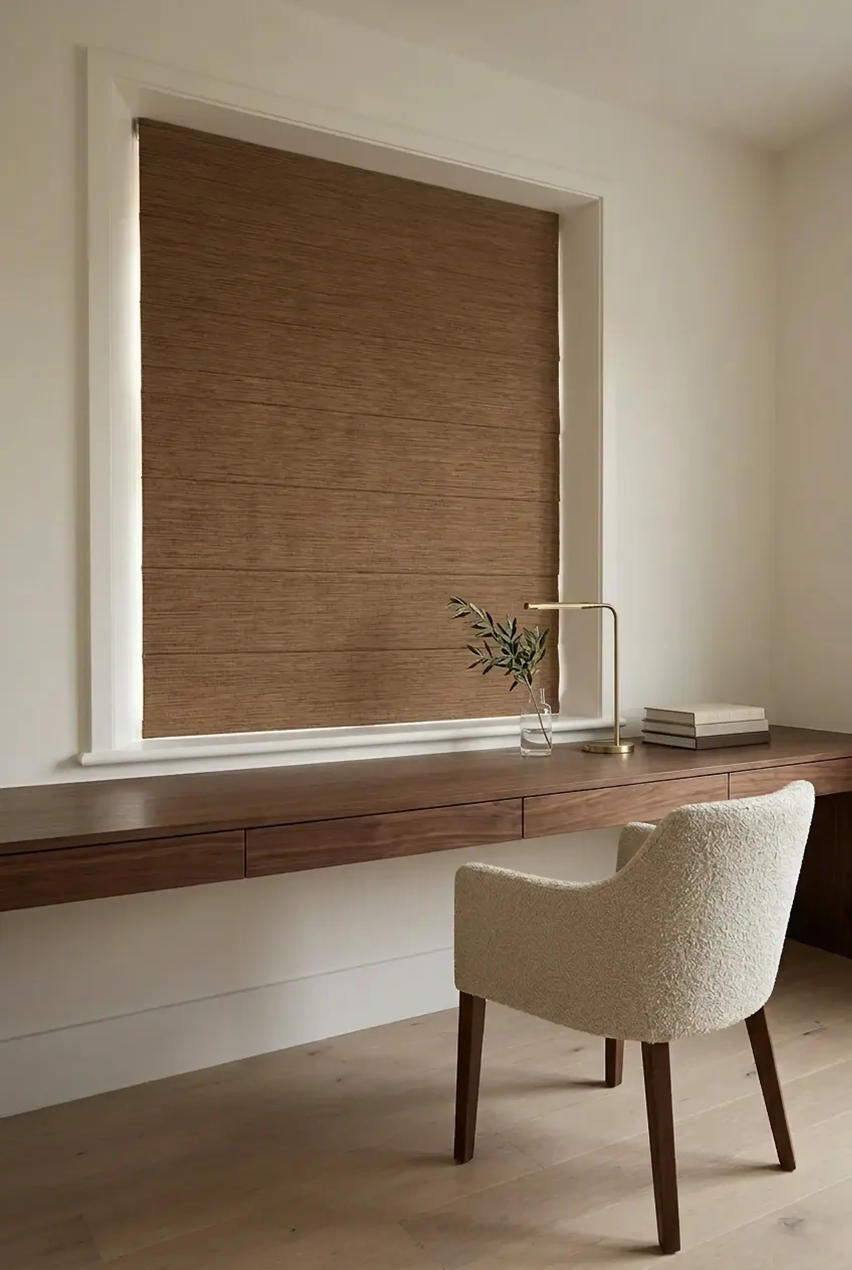 Brown woven Roman shade lowered inside a white window recess above a walnut desk with a bouclé chair, brass lamp and books in a calm UK home study