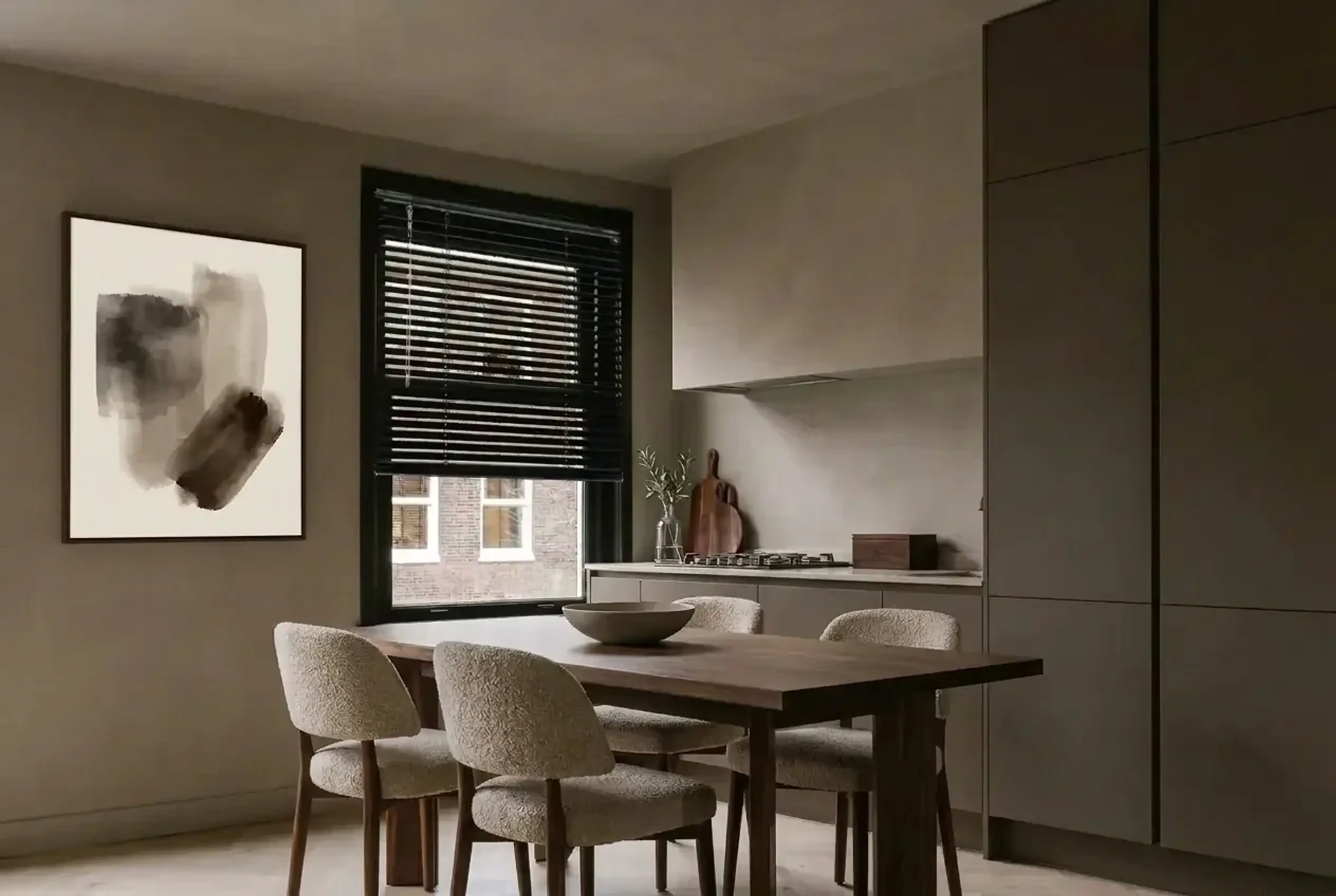 Black aluminium venetian blind on a black-framed window in a modern taupe kitchen with handleless units, a walnut dining table and bouclé dining chairs.
