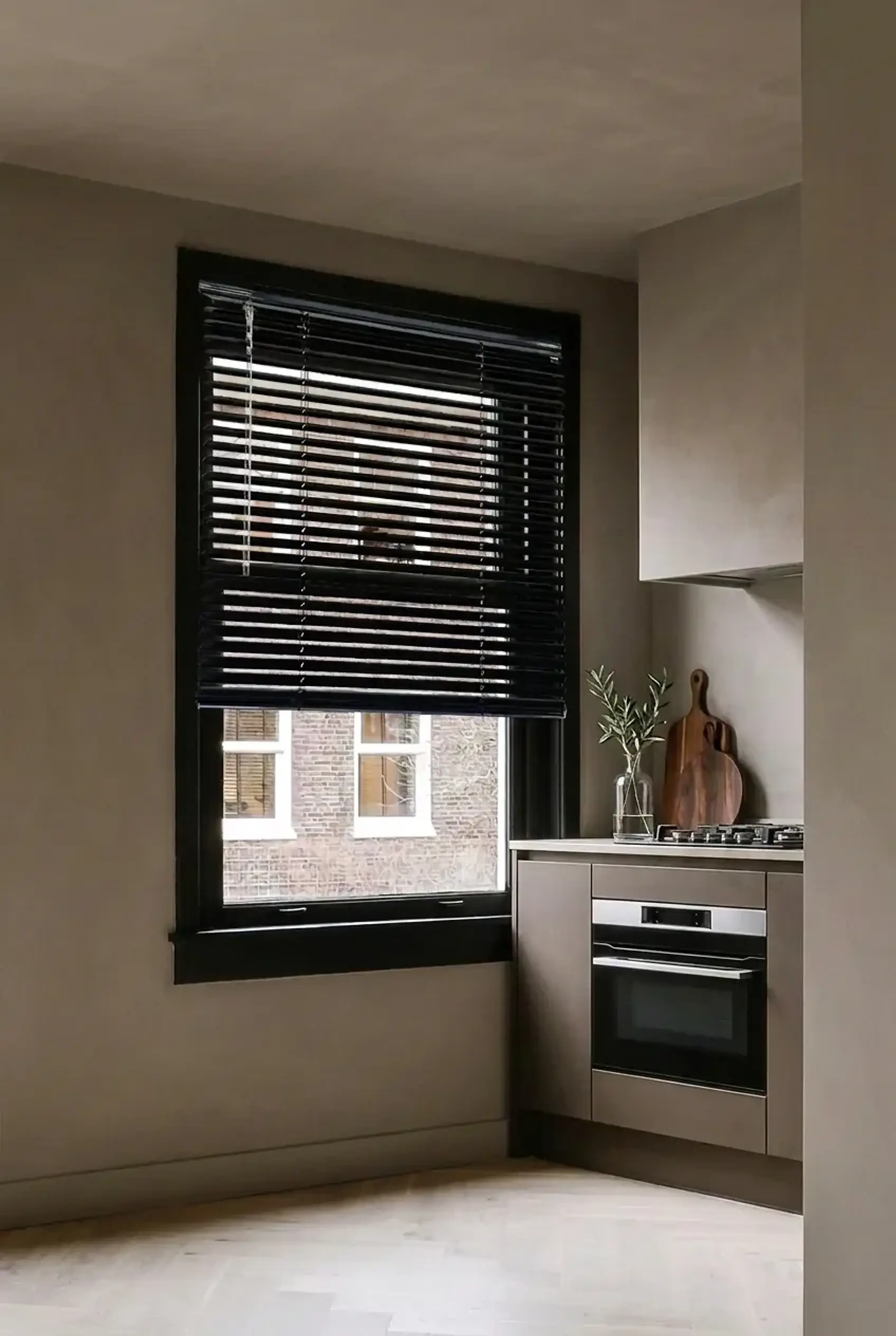 Black aluminium venetian blinds, partly raised, on a kitchen window with a black frame, next to matt mushroom-coloured cabinets and a stone worktop.