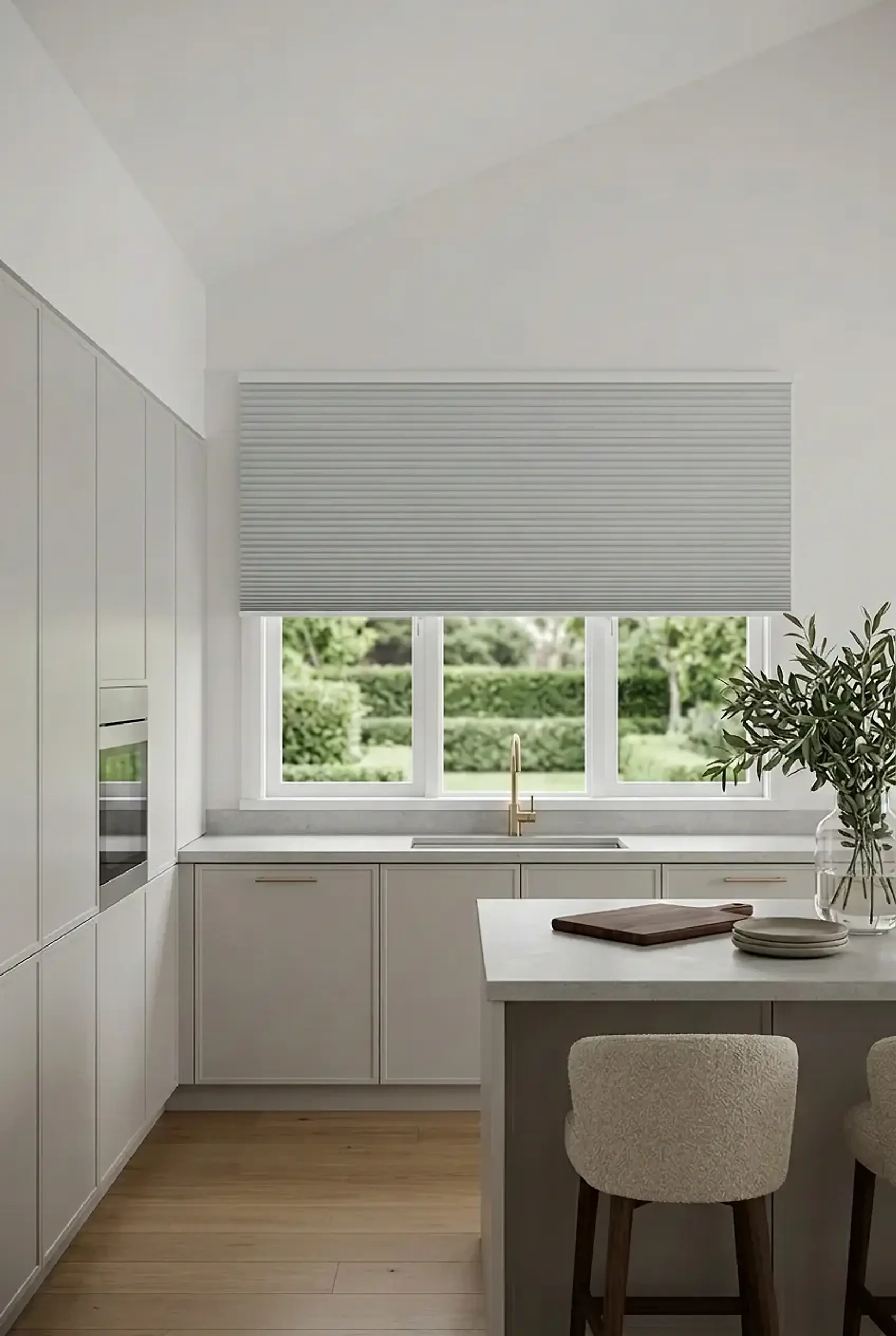 Pale grey honeycomb cellular shade above a kitchen window overlooking a green garden, with soft grey cabinetry, a brass tap and bouclé counter stools.