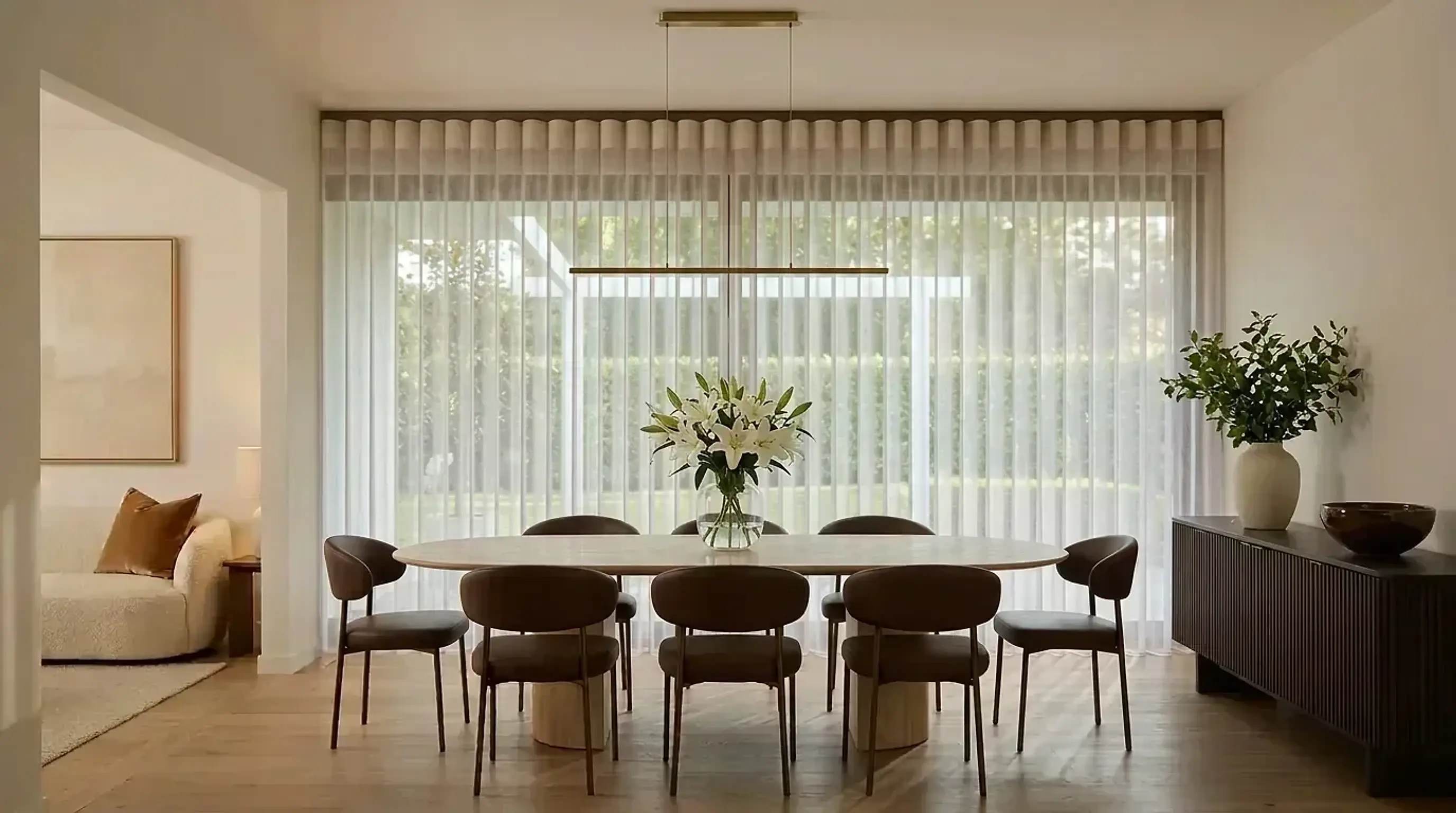 Floor-length SmartDrape sheer vanes across a glazed wall in an elegant dining room with a travertine table, chocolate leather chairs and white lilies.