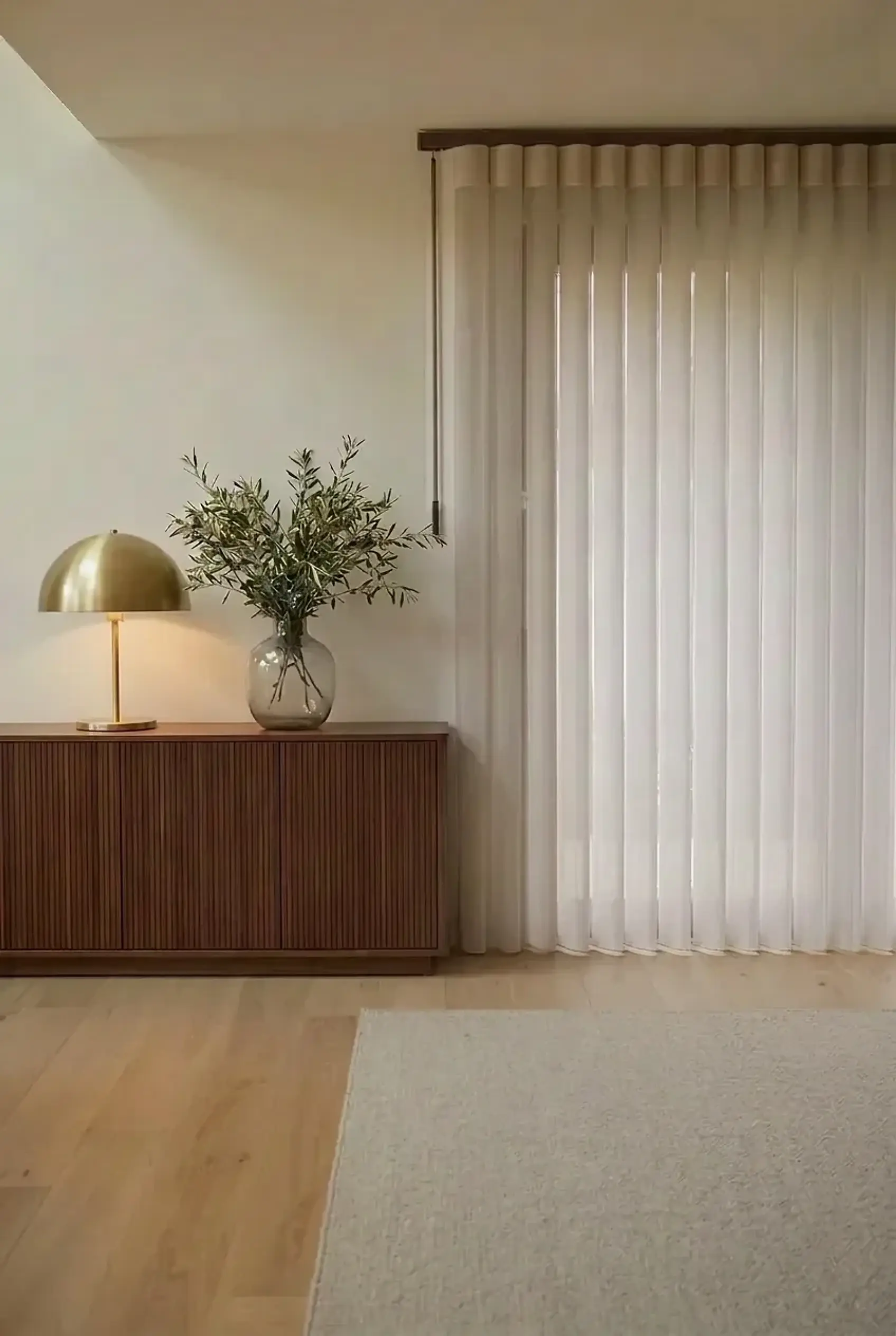 Sheer off-white SmartDrape vertical shade closed across a full-height glass door, the vanes falling to the floor, beside a fluted walnut sideboard with a brass lamp and olive branches in a calm UK home
