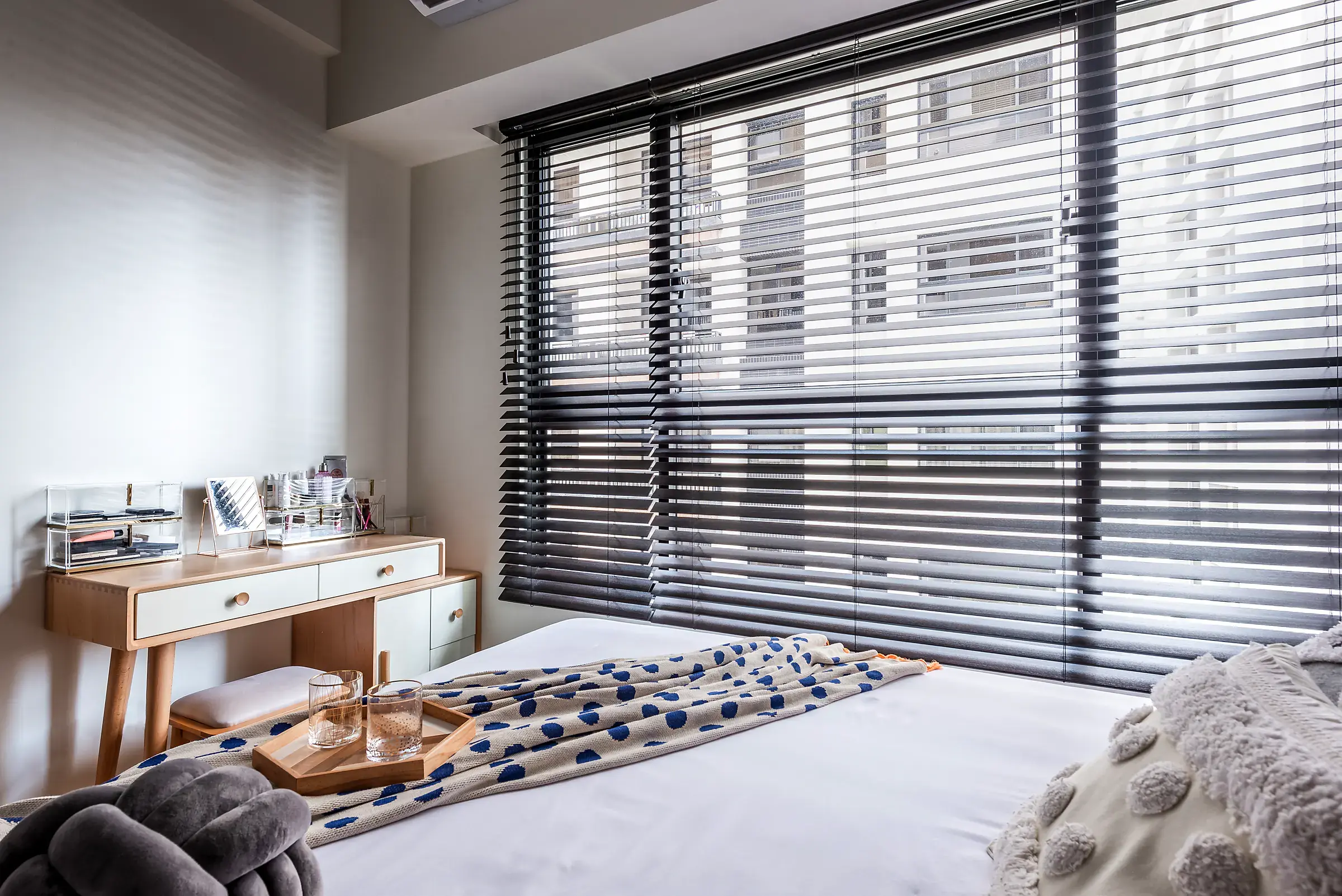 Black timber venetian blind across a wide bedroom window, slats tilted open to let in daylight over a white bed and a wooden dressing table.