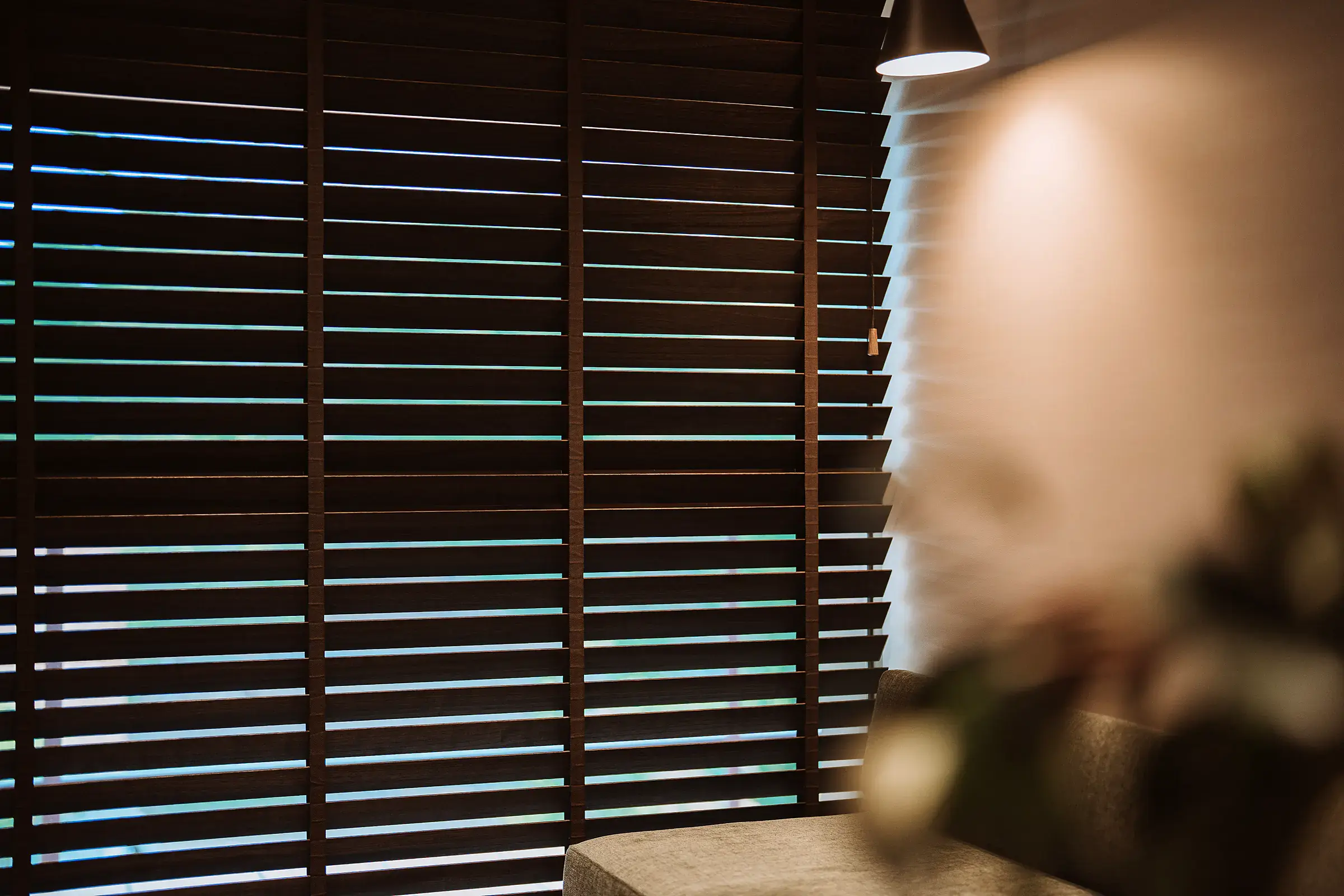Close-up of dark brown timber venetian blinds with matching ladder tapes, slats tilted to let slim lines of daylight through beside a grey sofa and a pendant light.