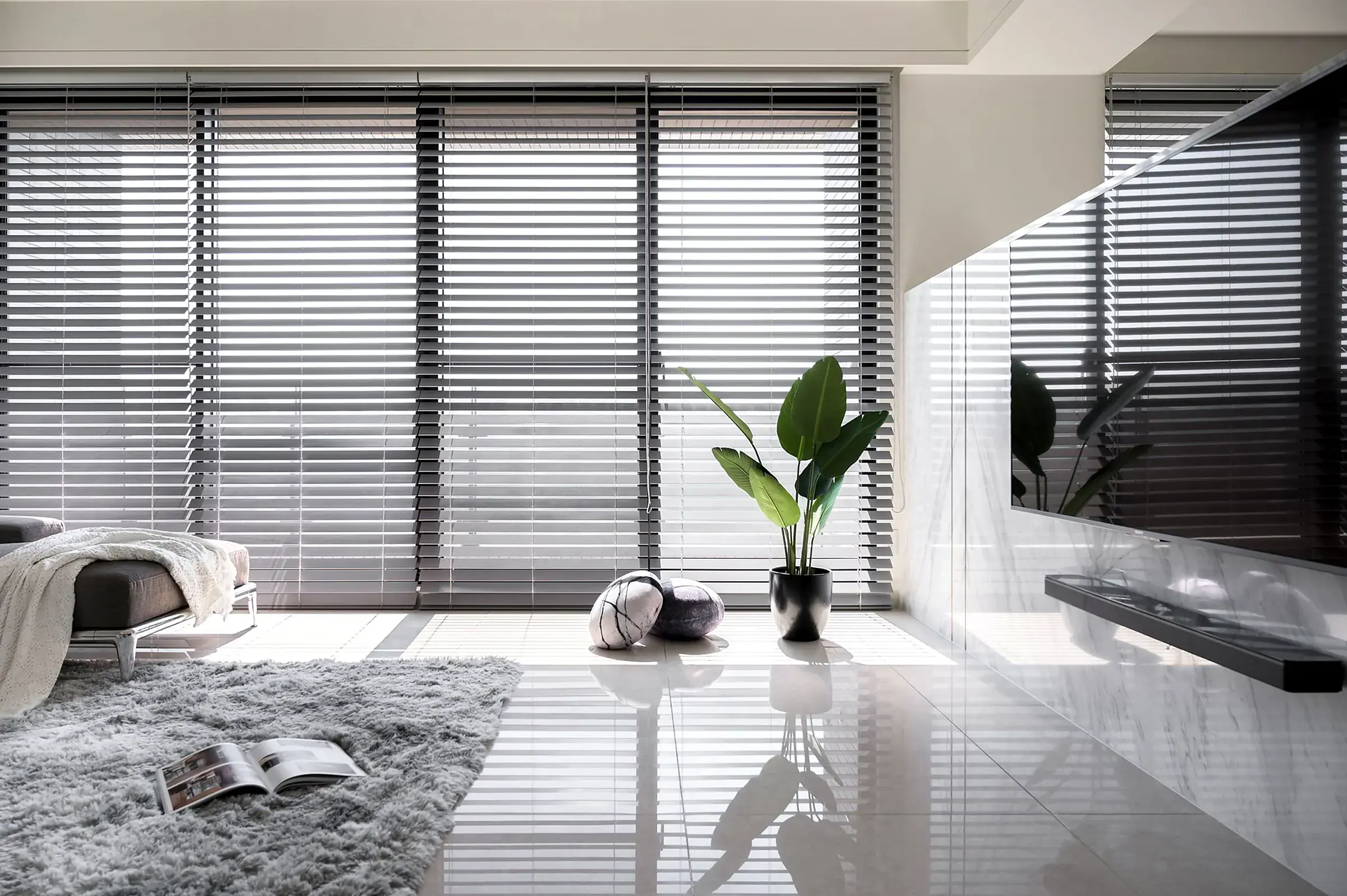 Grey timber venetian blinds across a wall of floor-to-ceiling windows in a bright living room, with slats tilted to let soft light onto a polished white floor.