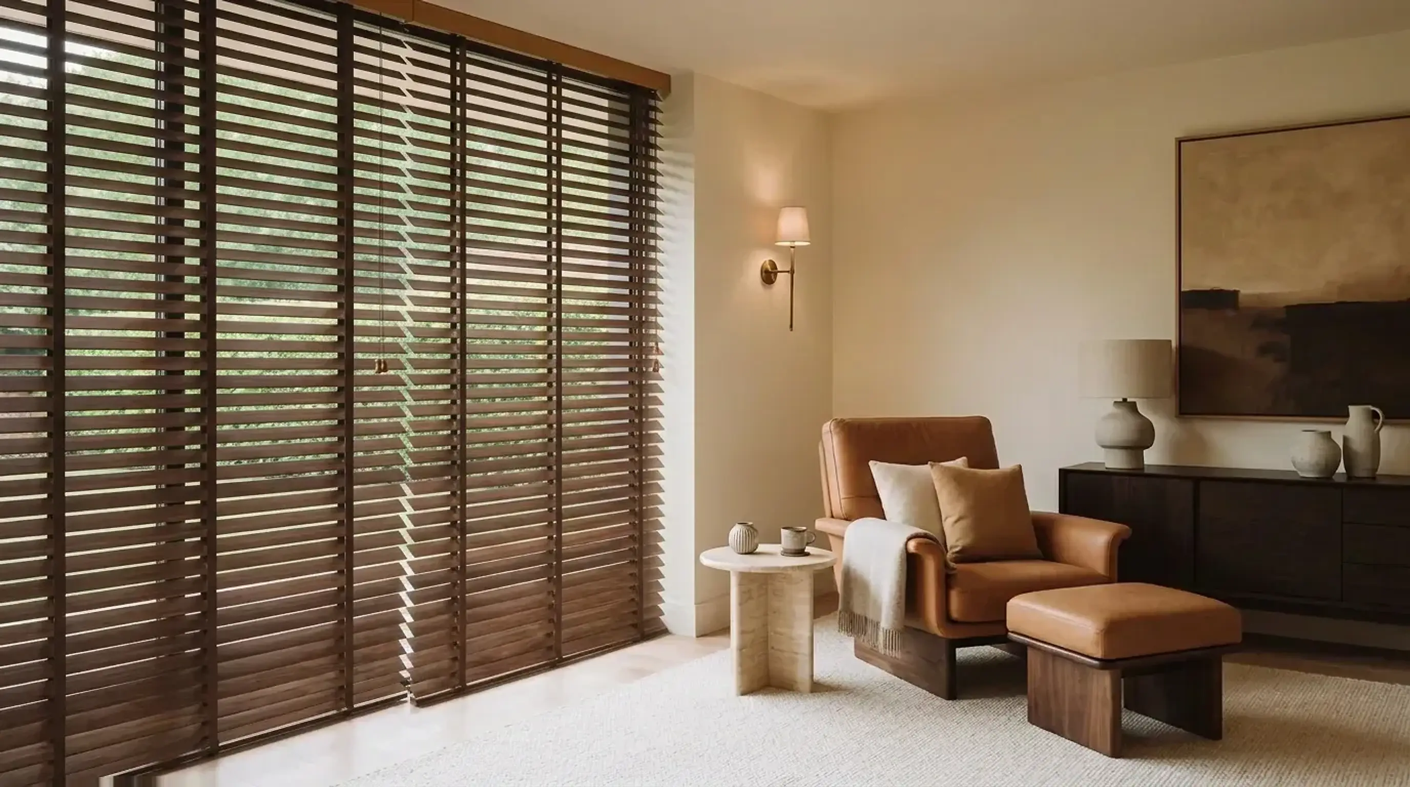 Two walnut timber venetian blinds with wide slats across a tall window in a warm ivory reading corner, with a cognac leather armchair and footstool and a travertine side table.