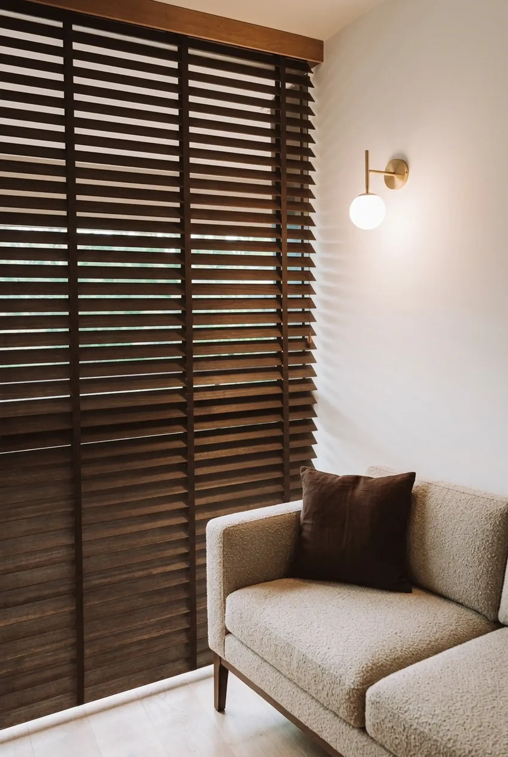Walnut timber venetian blinds with a matching timber pelmet in a warm-white living room, beside a modern oatmeal bouclé sofa and a brass wall light.