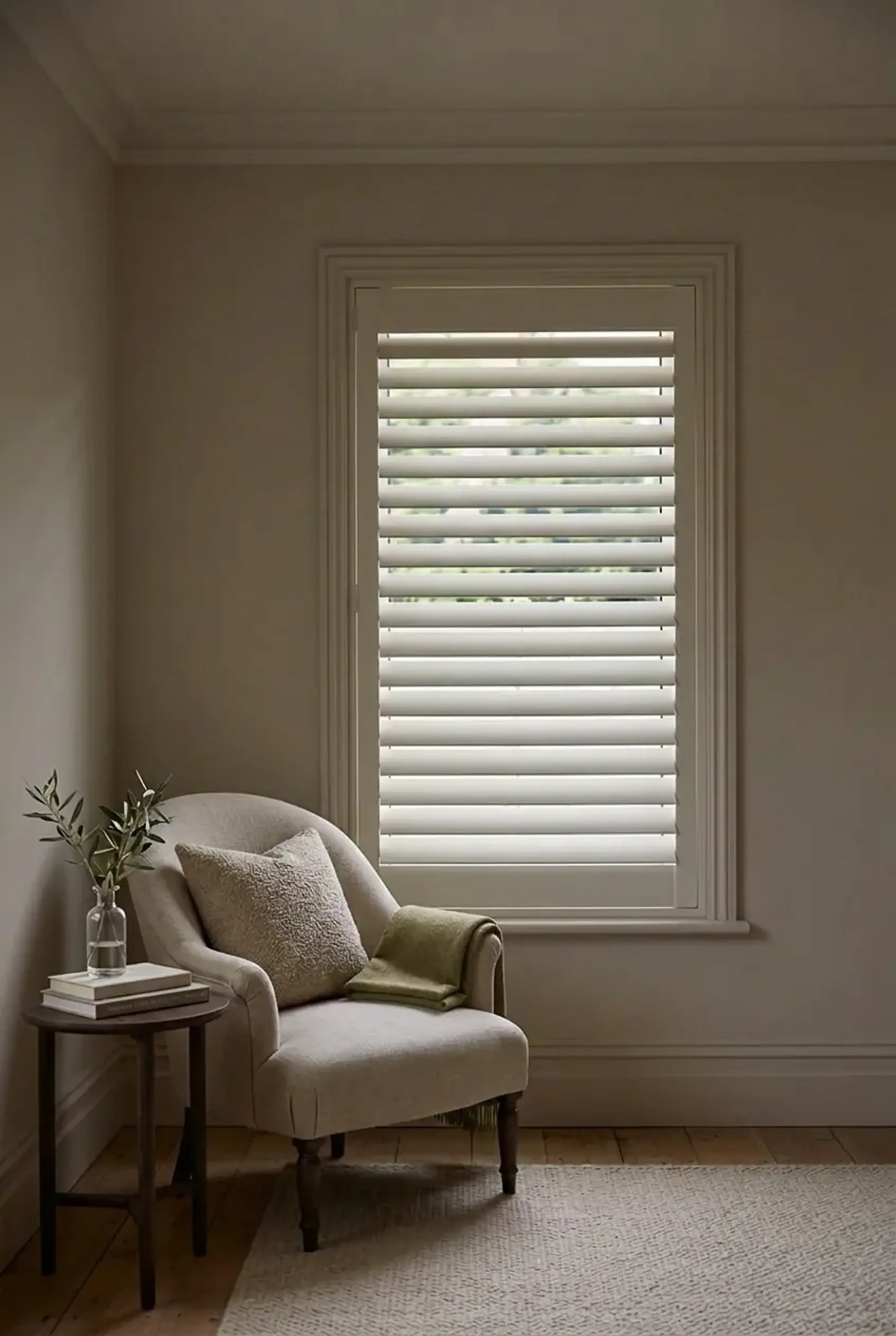 White Woodlore plantation shutters with wide louvres on a window in a calm, neutral English living room with a linen armchair.
